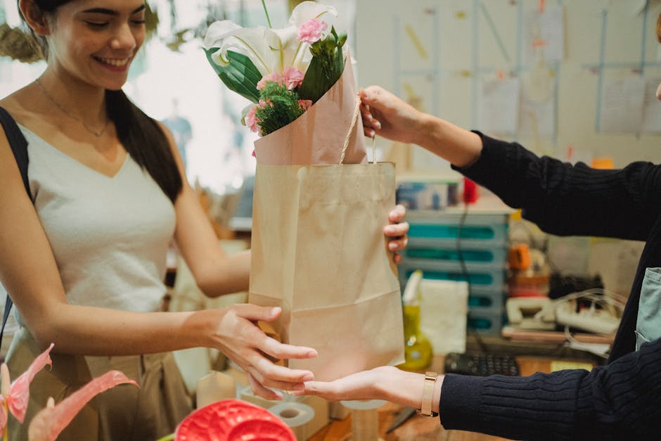 A woman with long dark hair, wearing a white sleeveless top, is smiling as she receives a bouquet of fresh flowers arranged with white lilies and pink roses, wrapped in pink and kraft paper, from a pe