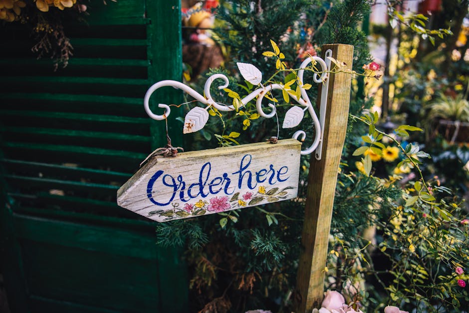A rustic wooden garden sign with the words 'Order here' painted in blue cursive, hanging from a white wrought iron bracket amid lush green foliage and flowering plants, with pink and yellow blooms vis
