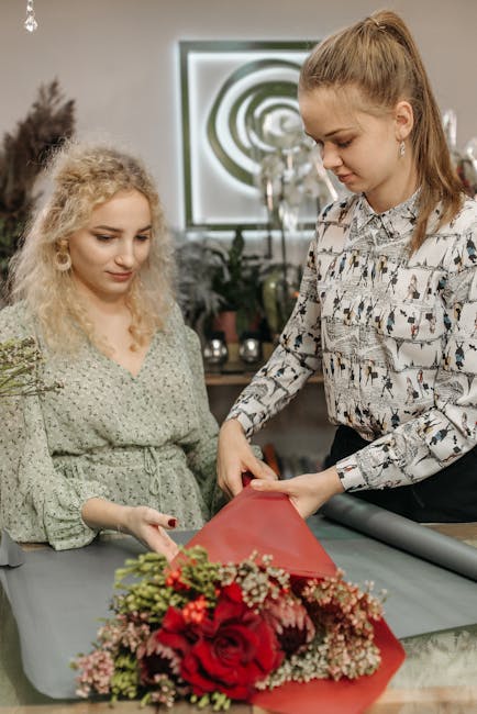 In a bright floral workshop, two women are engaged in creating a floral gift. The older woman, with long hair tied back and wearing a patterned blouse, holds a partially wrapped bouquet, while the you