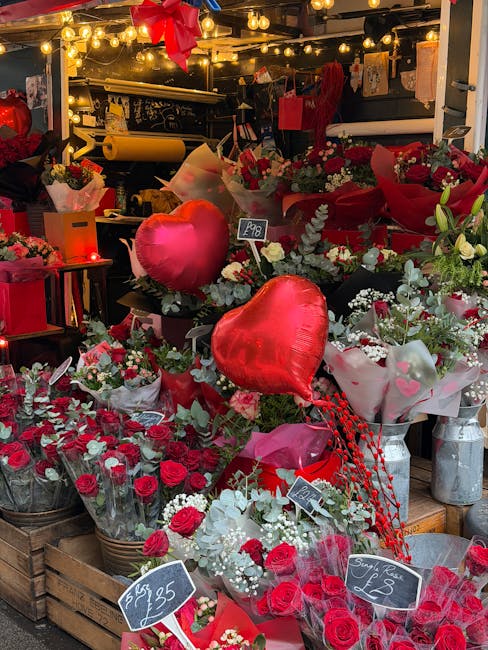 A vibrant floral display at a flower shop, featuring an assortment of fresh red roses, white baby's breath, and greenery arranged in bouquets. Bright red heart-shaped balloons are integrated into the 