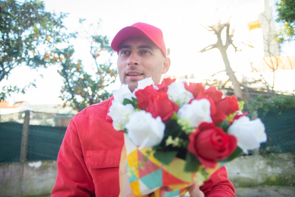 A man dressed in a red jacket and matching red cap is holding a colorful bouquet of fresh flowers, featuring red and white blooms with green foliage, wrapped in patterned paper. He stands outdoors in