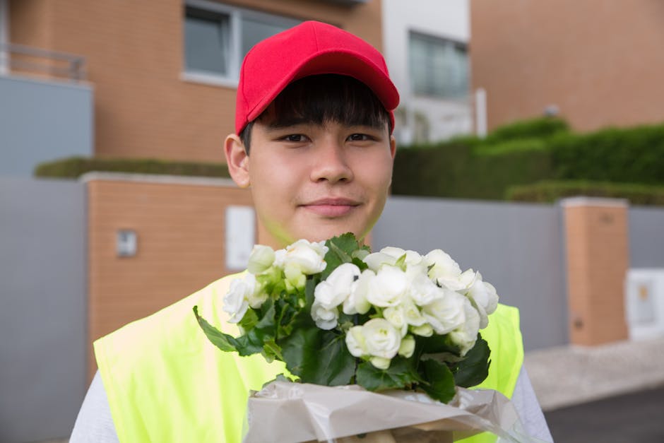 A young male flower delivery courier with a friendly expression, wearing a red cap and a high-visibility yellow vest, holding a bouquet of fresh white roses wrapped in tissue paper. He is positioned o