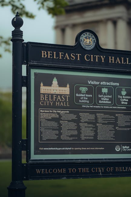 The image shows a black information signboard for Belfast City Hall with the text 'Belfast City Hall' and various visitor attraction details, set against a backdrop of trees and a cloudy sky. The sign