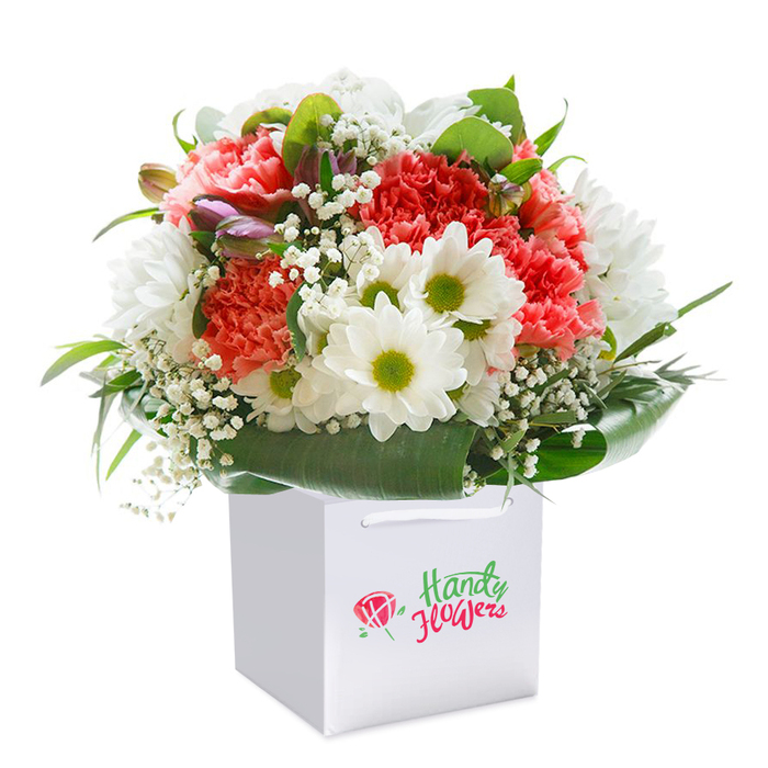 A breathtaking floral creation is arranged in a perfect, rounded bouquet, standing tall within a spotless, square white gift bag-its elegant presentation ideally suited for the classic surrounds of Harrow on the Hill. The composition is centred on an array of lush peach and coral carnations, their frilled and ruffled petals exuding texture and warmth, complemented by pristine white daisies that catch the eye with their bold, lime-yellow centres. Dewy sprigs of baby's breath are delicately woven throughout, lending an airy lightness and echoing the fresh scents one might encounter on a breezy day atop the hill or in Churchfields Park. Purple-tipped buds-partially closed and hinting at their coming bloom-add intrigue along the left, while a generous collar of deep, glossy foliage anchors the display, infusing depth and structure with every leaf. The white gift bag, accented by sturdy cord handles, ensures the bouquet is portable and presentation-ready for delivery to homes or venues near the High Street. Clear daylight gently illuminates the arrangement, enhancing every vermilion, peach, and creamy-white nuance for a look of pure, expressive abundance. Seamlessly blending local Harrow on the Hill spirit with sophisticated floral artistry, this arrangement is ideal for milestone celebrations, thoughtful gestures, or simply sharing nature's elegance, crafted with the signature skill and intuition of a neighbourhood florist.